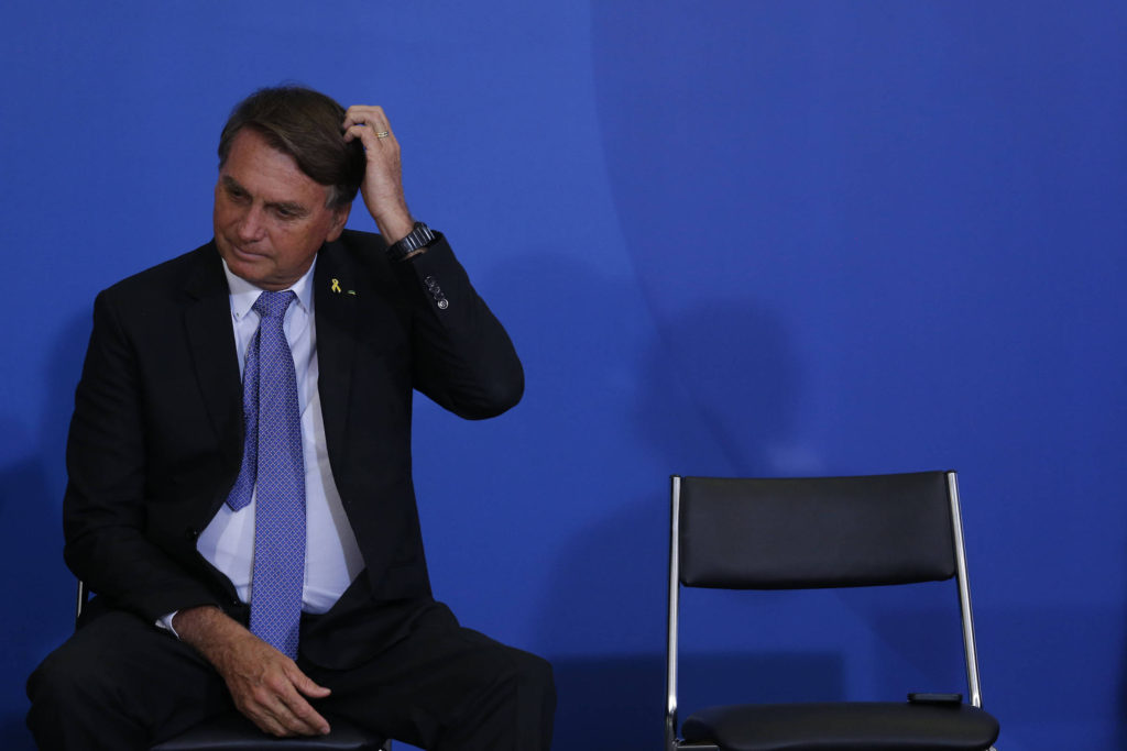 Foto de Jair Bolsonaro coçando a cabeça. Ao seu lado há uma cadeira preta vazia. A parede ao fundo é azul escuro.