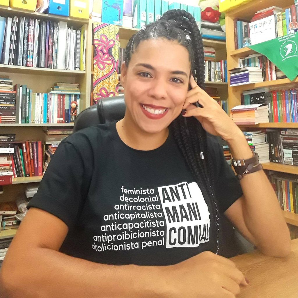 #ParaTodesVerem: retrato de Rachel Gouveia, ao fundo uma biblioteca. Ela veste uma camisa preta com os escritos estampados: ANTIMANICOMIAL. feminista, decolonial, antirracista, anticapitalista, anticapacitista, antiproibicionista e abolicionista penal. Crédito: Arquivo pessoal/Rachel Gouveia